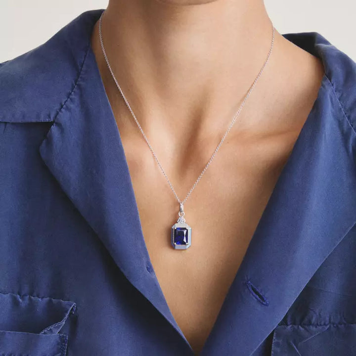 A close-up of a model wearing a silver chain necklace with a rectangular blue gemstone pendant, styled with a blue satin shirt.