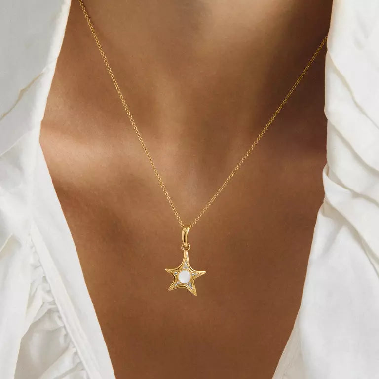 A gold star-shaped pendant necklace with a clear central gemstone, worn on a model's neck over a white blouse.