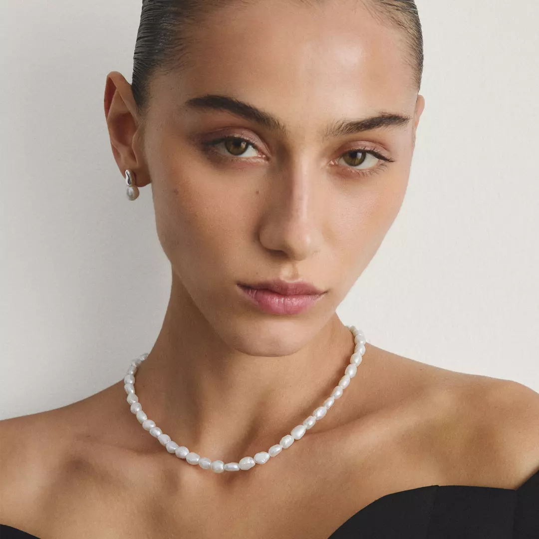 A close-up portrait of a model wearing a delicate white pearl necklace and a matching small pearl stud earring against a soft light gray background.