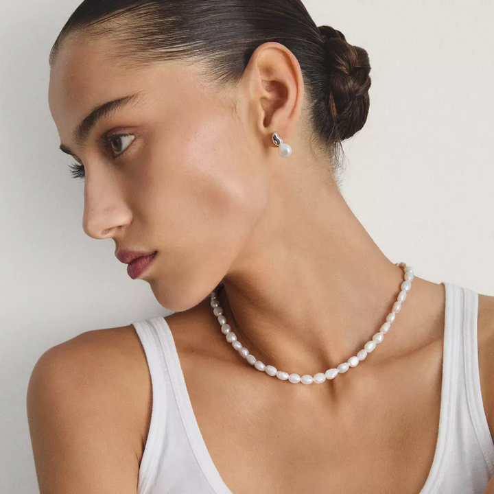 A woman with her hair in a bun wears a white tank top, a white pearl necklace, and a small white pearl stud earring against a plain light background.