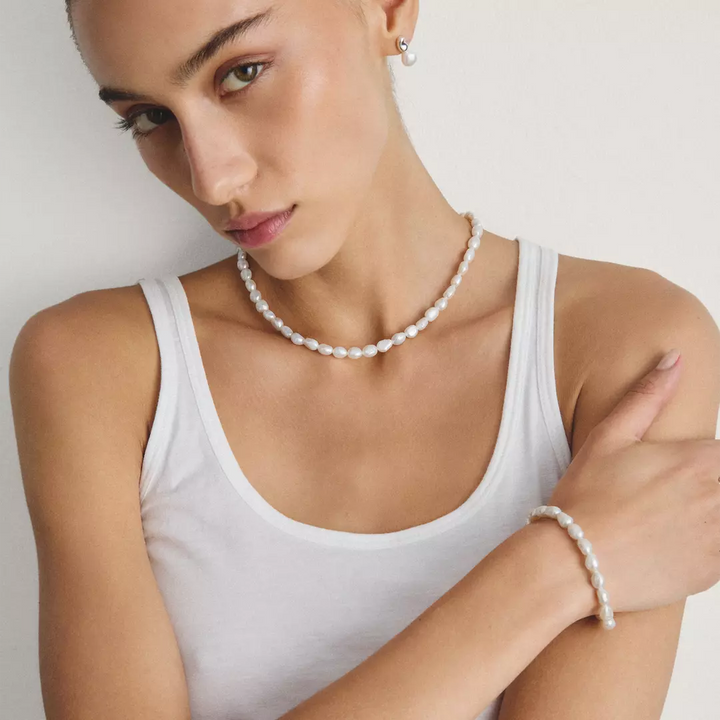 A woman wearing a white ribbed tank top models a white pearl necklace and matching pearl bracelet against a clean, off-white background.