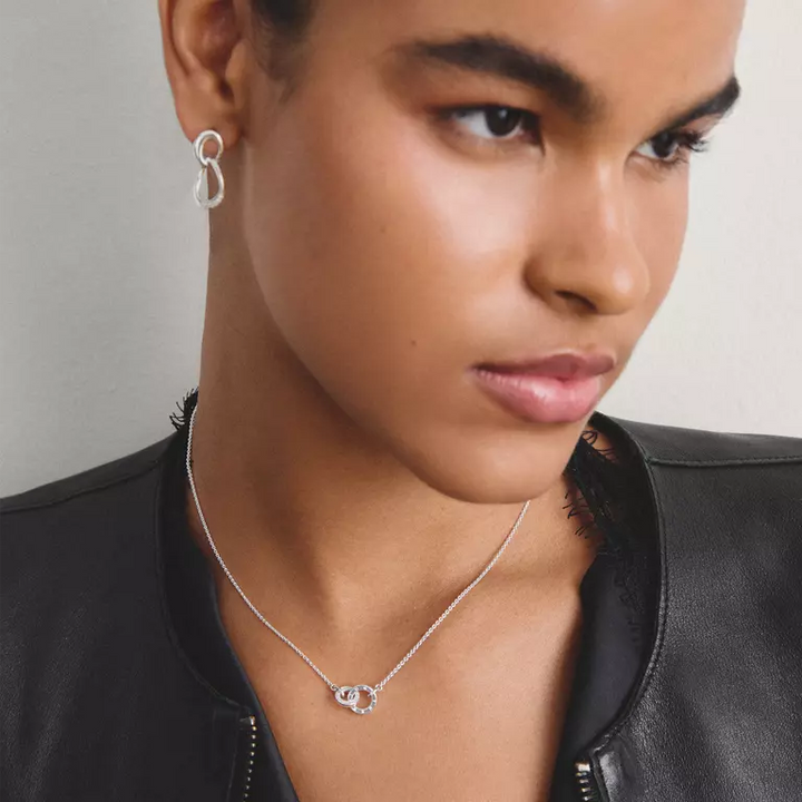 A close-up of a model wearing a silver infinity-shaped pendant necklace and a matching silver hoop earring, styled with a black leather jacket against a neutral gray background.