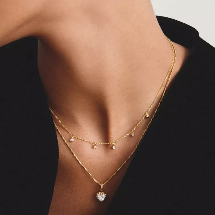 A close-up of a model's neck and upper chest wearing two layered gold necklaces. The shorter necklace features small dangling clear stones, while the longer necklace has a heart-shaped clear pendant at the bottom.