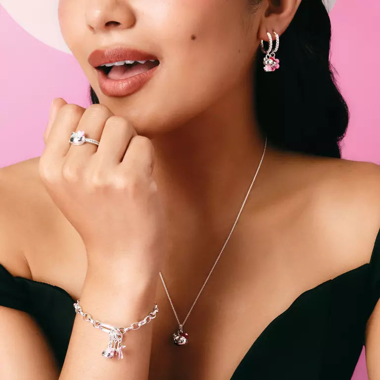 A close-up lifestyle shot of a woman wearing a matching silver jewelry set, including a ring, necklace, and bracelet, along with matching hoop earrings. The jewelry features clear crystals and pink gemstone accents, set against a solid pink background.