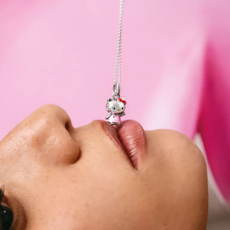 A close-up lifestyle shot of a person's lips touching a silver Hello Kitty pendant necklace hanging vertically against a soft pink background.