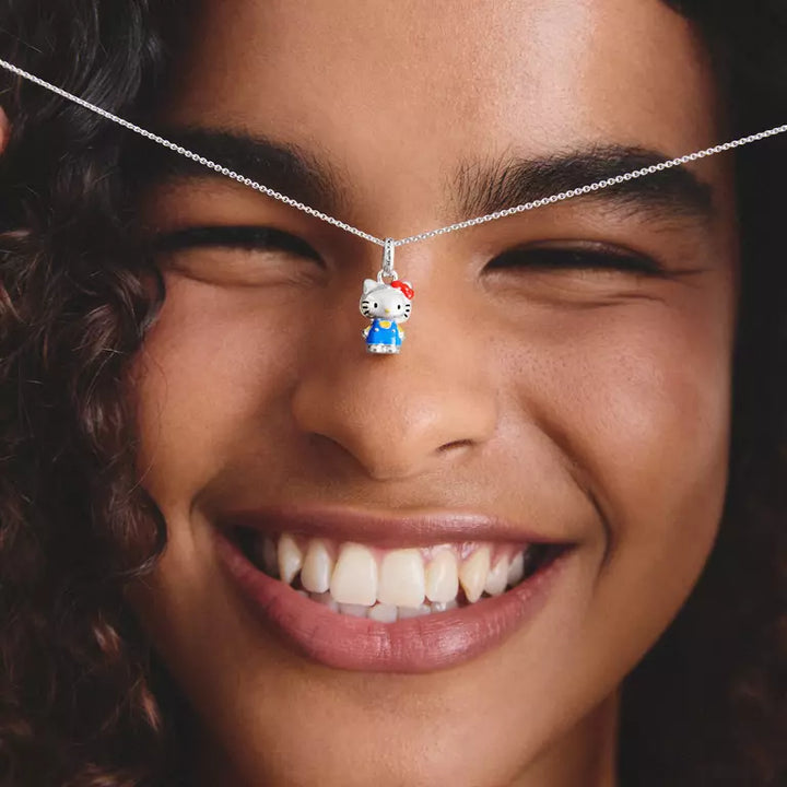 A close-up of a smiling woman wearing a delicate silver necklace with a small Hello Kitty pendant. The pendant features Hello Kitty's white face, red bow, and blue dress, suspended against her eyes.