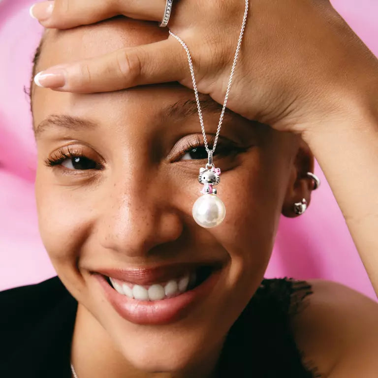 A close-up lifestyle shot of a smiling woman with short hair, wearing a silver pendant necklace with a white pearl and a rhinestone crown. She is holding her hand to her forehead, with a solid pink background.