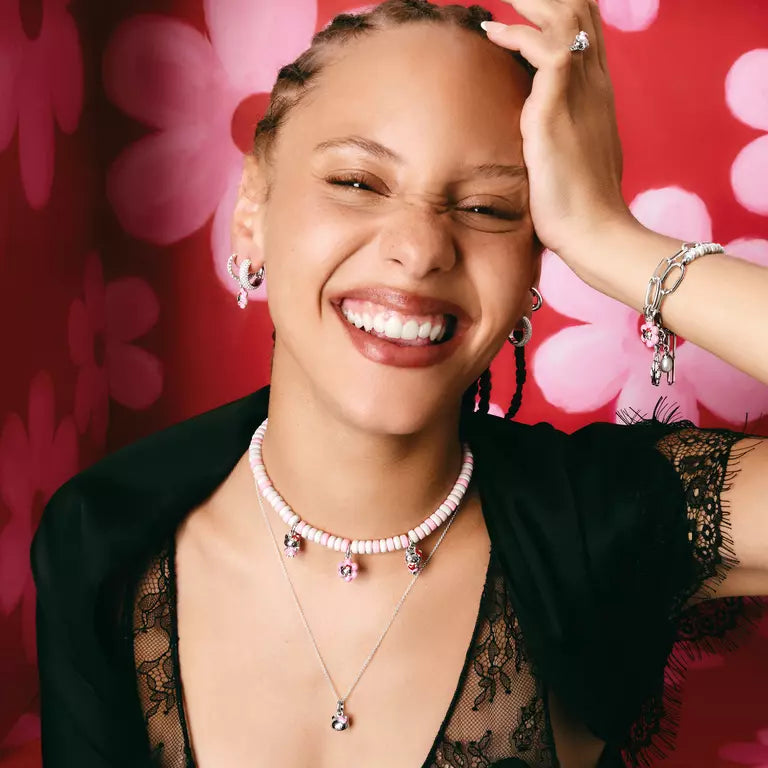 A smiling woman wearing multiple jewelry pieces, including layered necklaces, hoop earrings, bracelets, and a ring, posed against a bright red background with pink floral shapes.