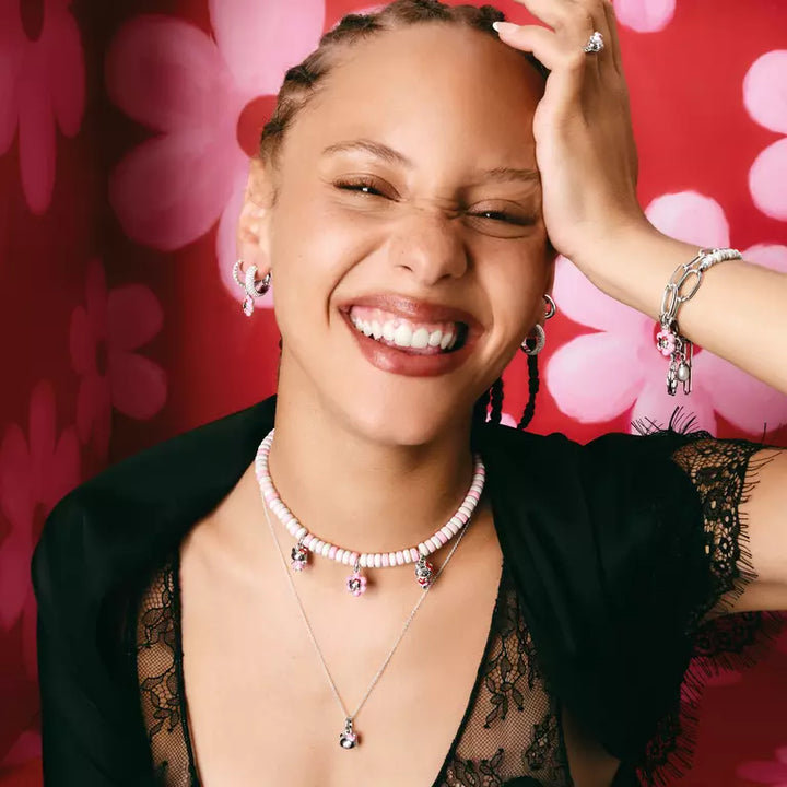 A smiling woman wearing multiple jewelry pieces, including layered necklaces, hoop earrings, bracelets, and a ring, posed against a bright red background with pink floral shapes.