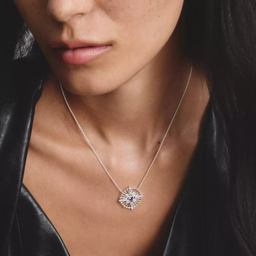 A close-up of a model wearing a delicate silver chain necklace with a sunburst pendant featuring a central blue gemstone.