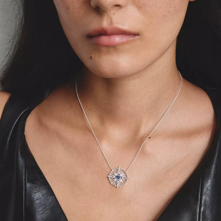 A close-up of a model wearing a delicate silver chain necklace with a starburst-shaped silver pendant featuring a central blue gemstone.