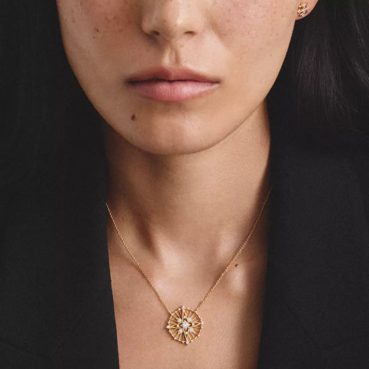 Close-up of a model wearing a delicate gold chain necklace with a sunburst flower pendant featuring white stones, paired with a matching gold stud earring.