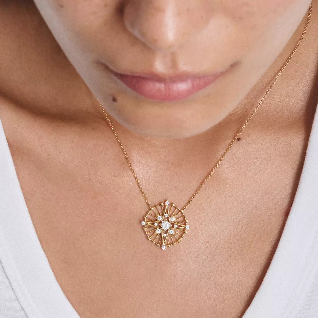 A close-up of a person wearing a delicate gold necklace with a geometric starburst pendant featuring small clear stones, shown against the wearer's neck and upper chest.