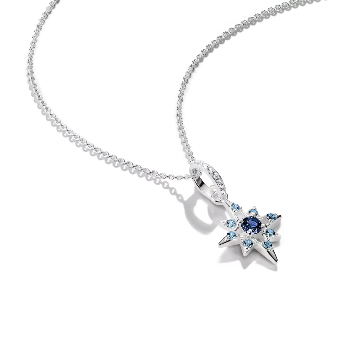 A silver-toned starburst pendant necklace featuring a central dark blue gemstone surrounded by small light blue and clear stones, suspended from a delicate silver chain on a white background.