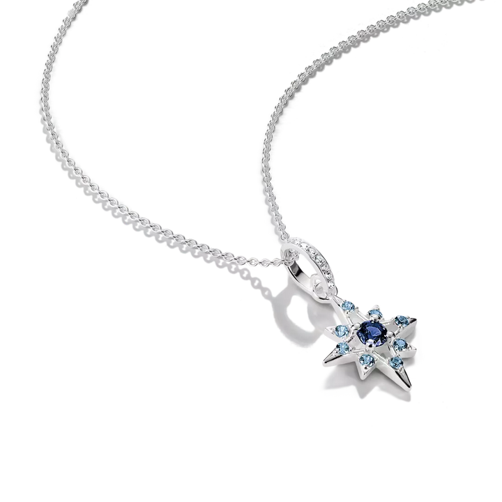 A silver-toned starburst pendant necklace featuring a central dark blue gemstone surrounded by small light blue and clear stones, suspended from a delicate silver chain on a white background.