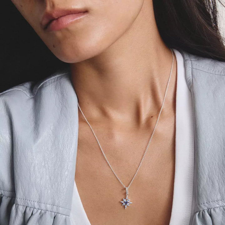 A silver-toned starburst pendant necklace featuring a central dark blue gemstone surrounded by small light blue and clear stones, suspended from a delicate silver chain on a white background.