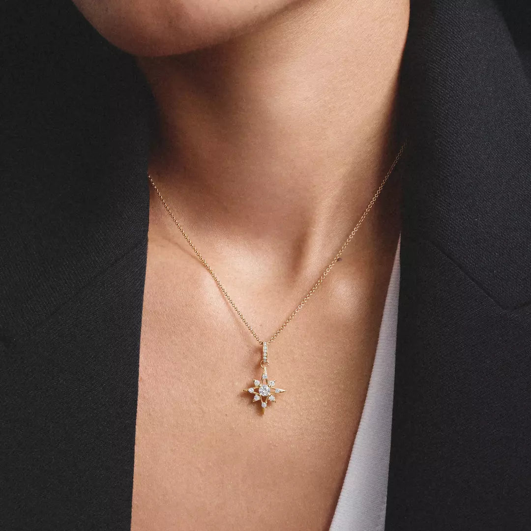 A close-up of a person wearing a delicate gold chain necklace with a small starburst pendant set with clear stones, styled with a black blazer.
