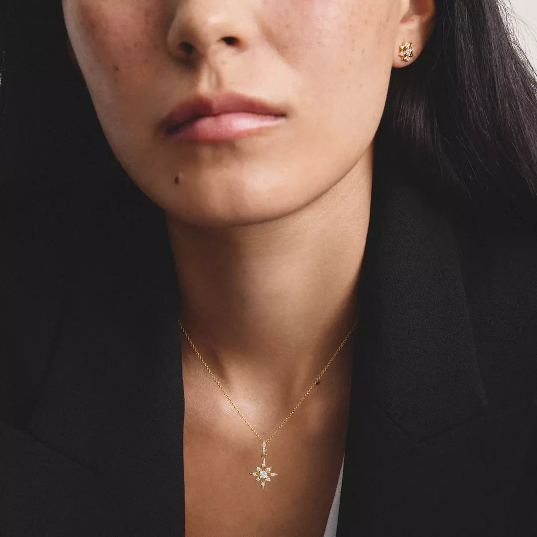 A close-up of a woman wearing a delicate gold necklace with a small snowflake or starburst pendant, along with matching gold stud earrings, styled with a black blazer.