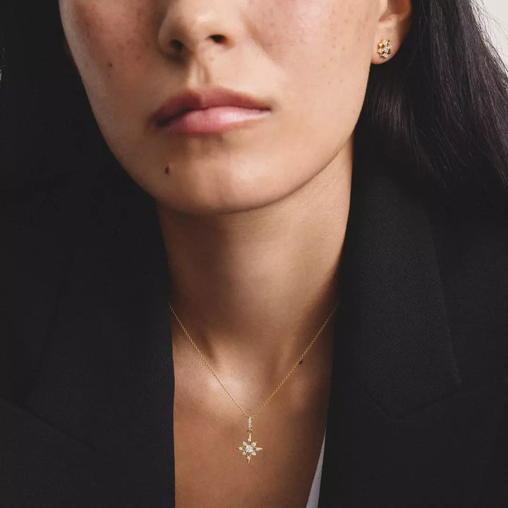 A close-up of a woman wearing a delicate gold necklace with a small snowflake or starburst pendant, along with matching gold stud earrings, styled with a black blazer.