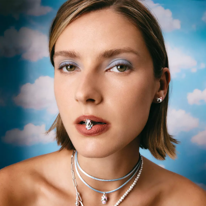 A close-up portrait of a woman with short blonde hair wearing multiple layered necklaces and a lip ring against a blue sky with clouds backdrop.