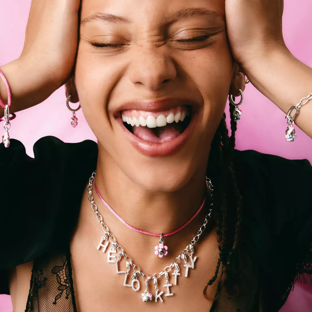 A close-up lifestyle shot of a smiling woman with eyes closed, wearing layered necklaces including a silver 'HELLO KITTY' letter necklace with pink rhinestones and a pink cord necklace, along with matching pink and silver bracelets, set against a solid pink background.