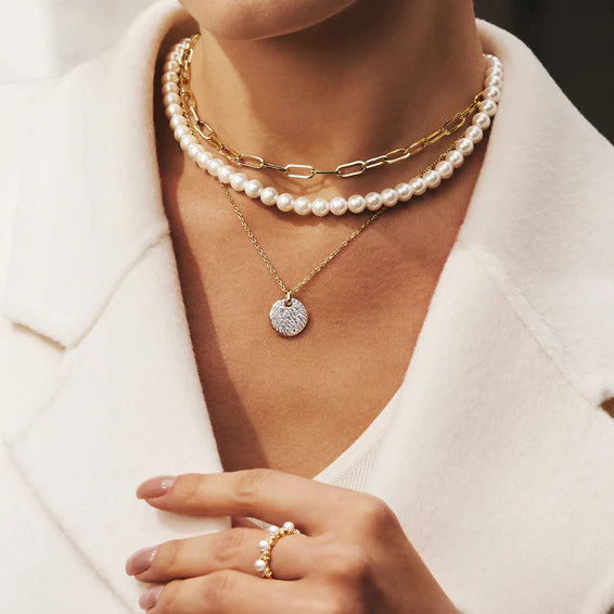 Person wearing a pearl necklace and gold chain with a close-up of a hand holding a ring.