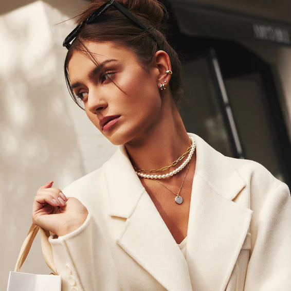Woman in a white coat with jewelry, holding a handbag, against a blurred background