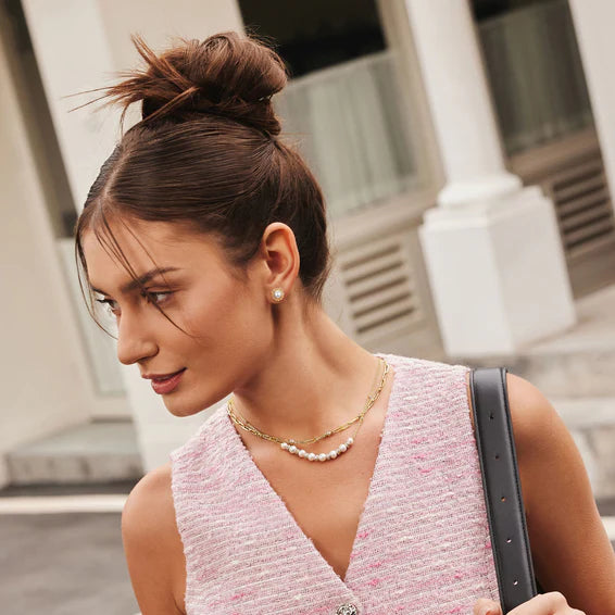 Woman in a pink textured dress with a gold necklace and pearl earrings, standing outdoors.