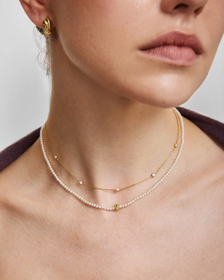 Close-up of a model wearing a double-layered necklace with one layer of small white pearls and one layer of delicate gold chain with tiny gold beads and a small gold charm, along with a gold hoop earring.