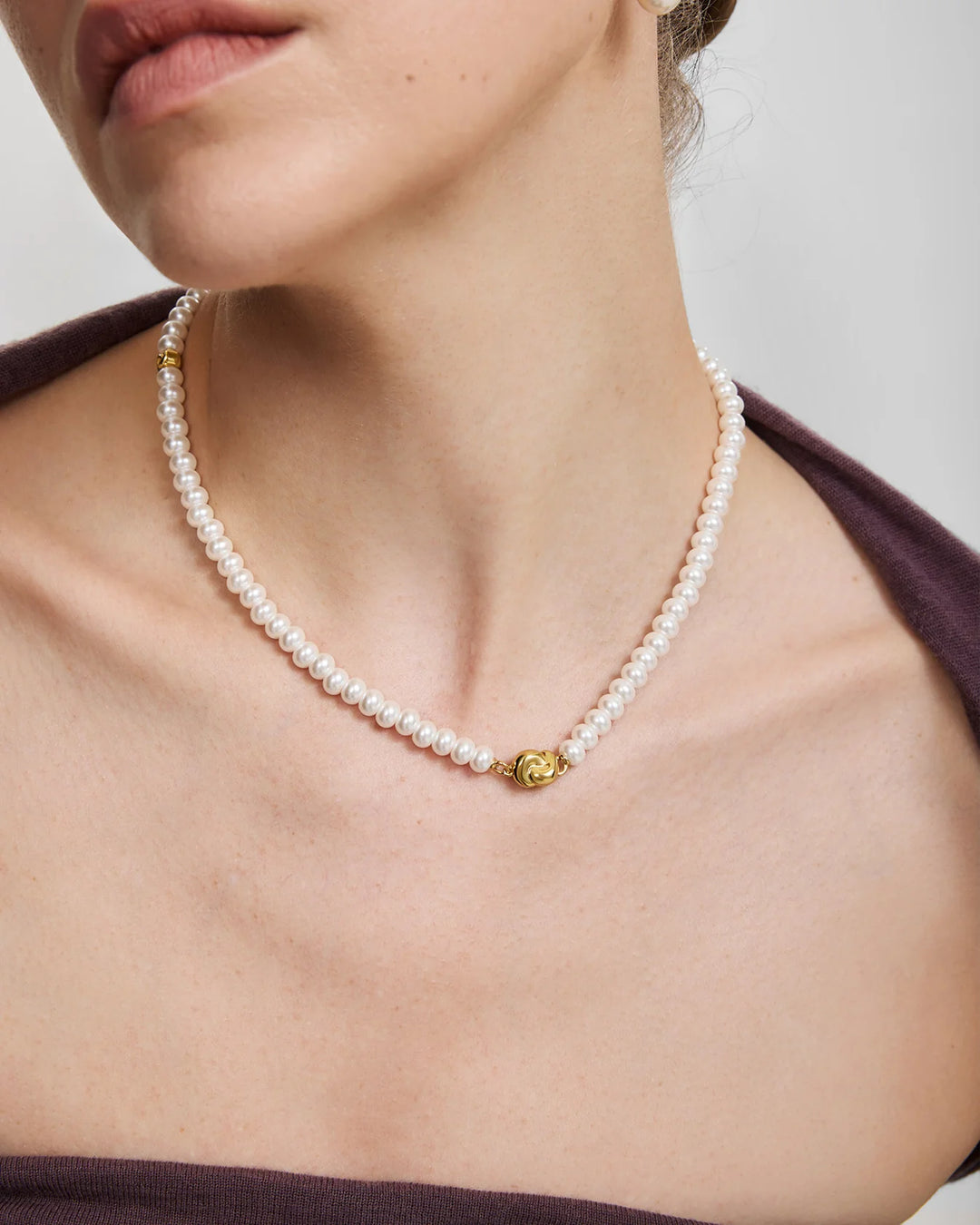 A close-up of a model wearing a delicate white pearl necklace with a small gold lion-head charm at the center, styled with a dark brown strap.