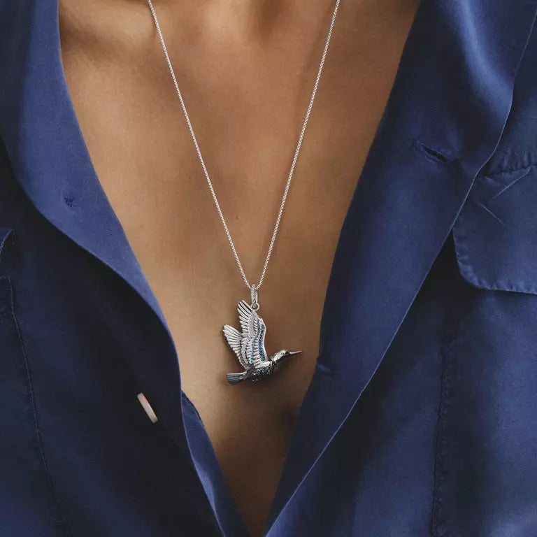 A silver bird pendant necklace worn by a model in a navy blue satin shirt. The necklace features a detailed flying bird pendant on a delicate silver chain.