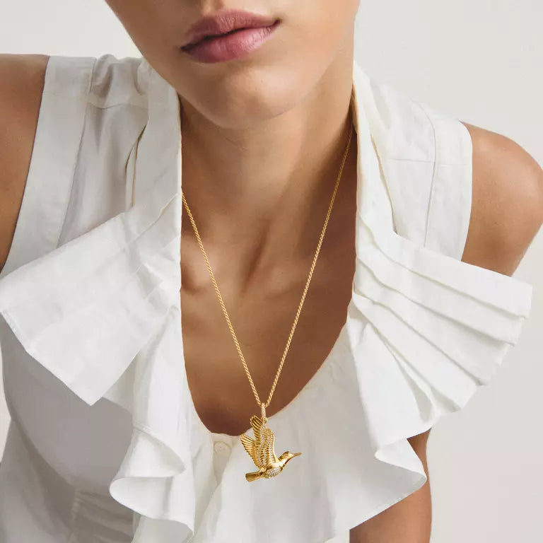 A close-up of a model wearing a delicate gold chain necklace with a small flying bird pendant, styled with a white ruffled top against a light neutral background.