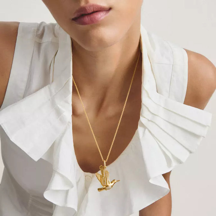 A close-up of a model wearing a delicate gold chain necklace with a small flying bird pendant, styled with a white ruffled top against a light neutral background.