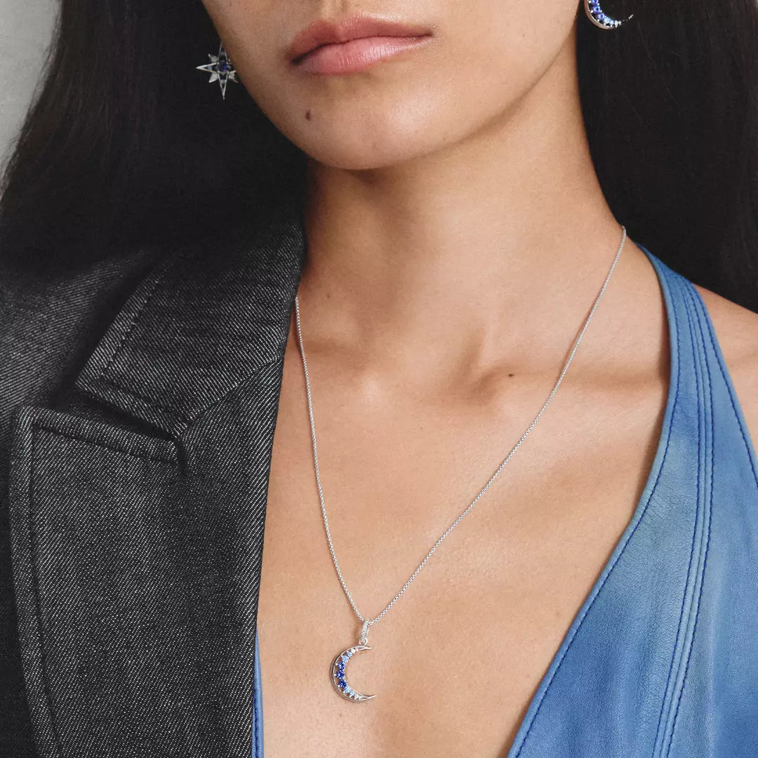 A close-up of a person wearing a silver crescent moon pendant necklace with blue and red gemstone accents, styled with a dark gray blazer.