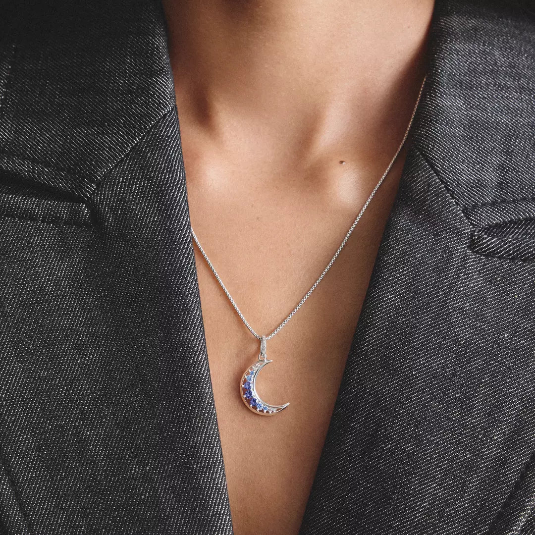 A close-up of a person wearing a silver crescent moon pendant necklace with blue and red gemstone accents, styled with a dark gray blazer.