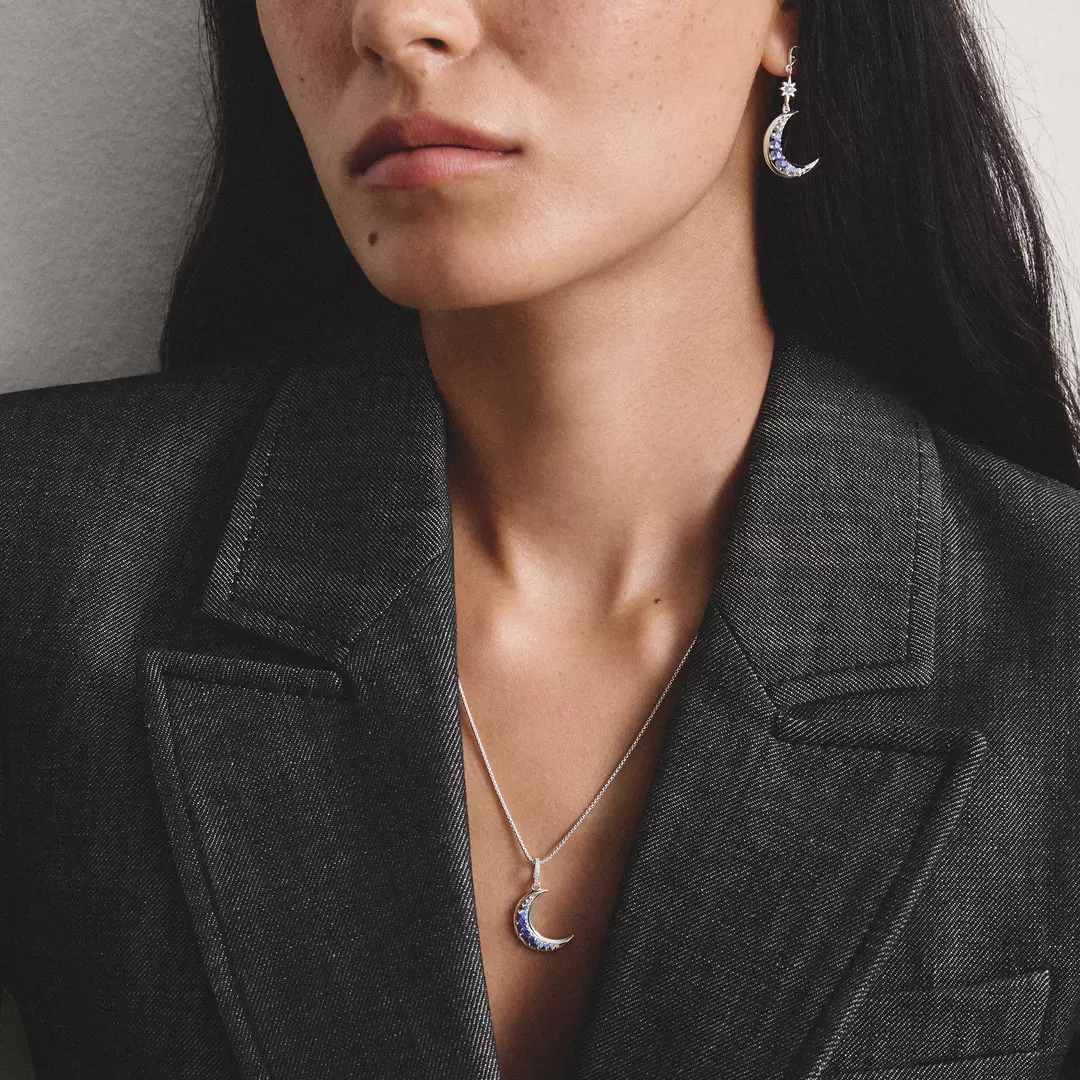 Close-up of a woman wearing a silver crescent moon pendant necklace and matching crescent drop earrings with small blue gemstone accents, styled with a dark gray blazer.