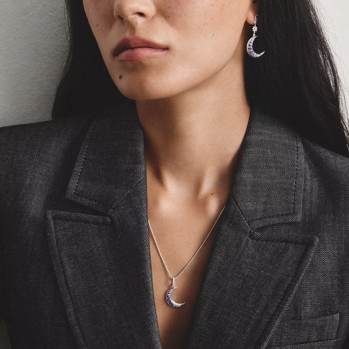 Close-up of a woman wearing a silver crescent moon pendant necklace and matching crescent drop earrings with small blue gemstone accents, styled with a dark gray blazer.