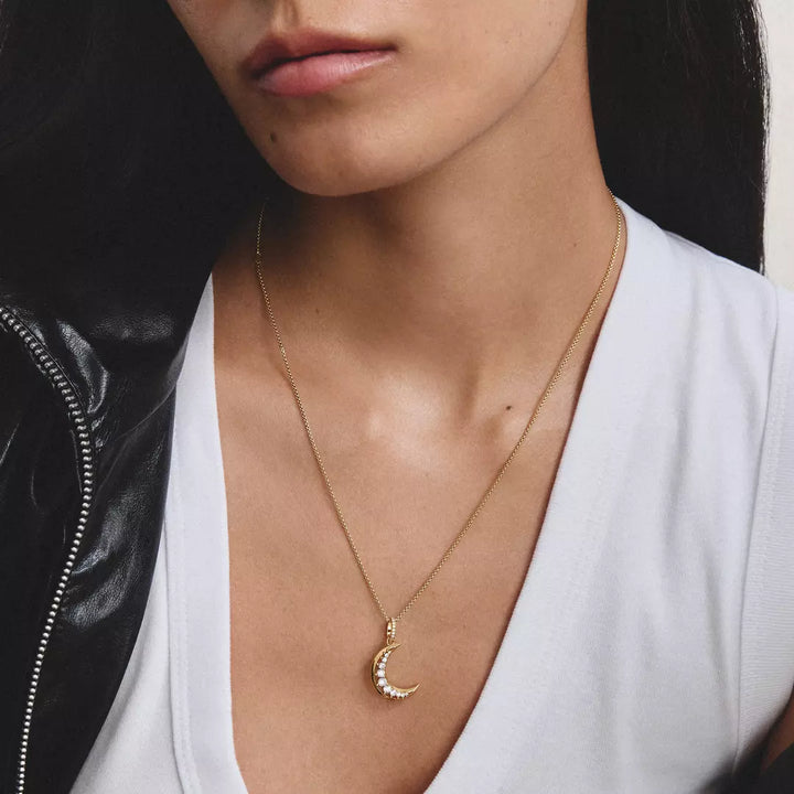 A close-up of a model wearing a delicate gold crescent moon pendant necklace with small clear stones along the edge, styled with a white top and a black leather jacket.