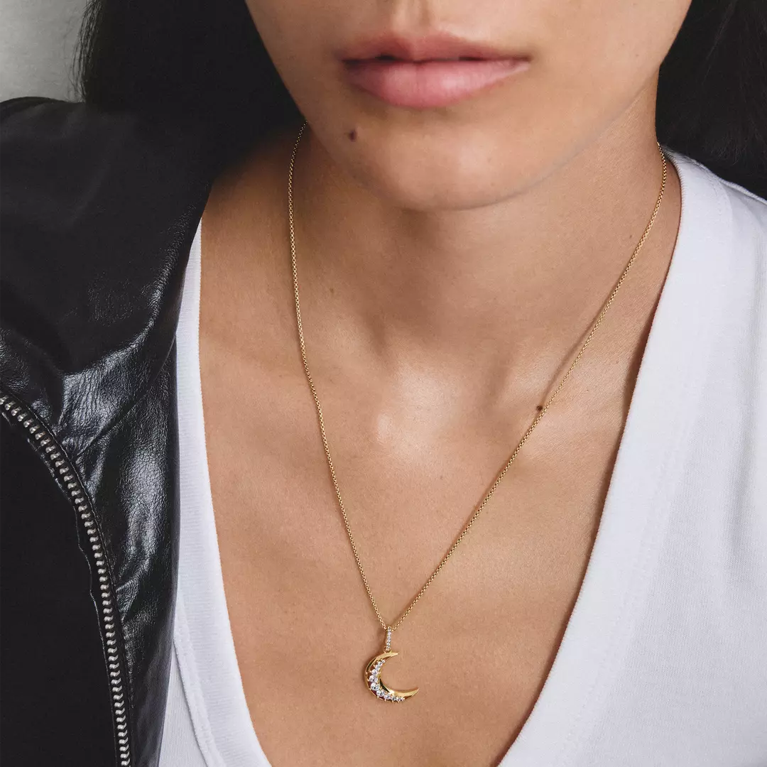 A close-up of a model wearing a delicate gold crescent moon pendant necklace with small clear stones along the edge, styled with a white top and a black leather jacket.