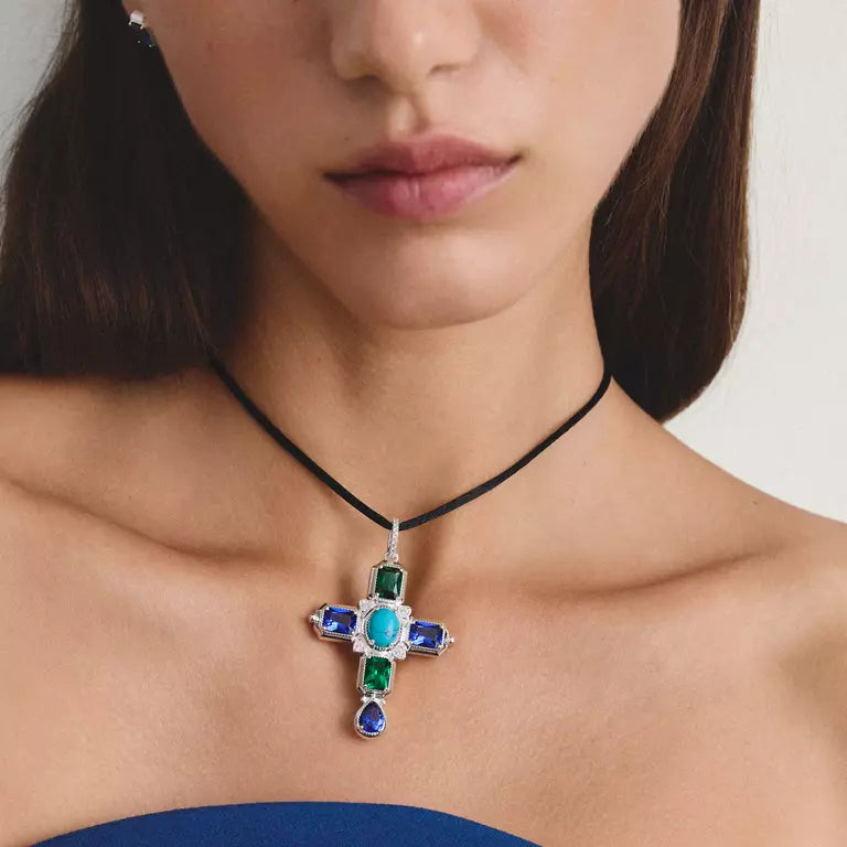 A close-up of a model wearing a silver cross pendant necklace with green, blue, and turquoise stones on a black cord, paired with a blue strapless top.