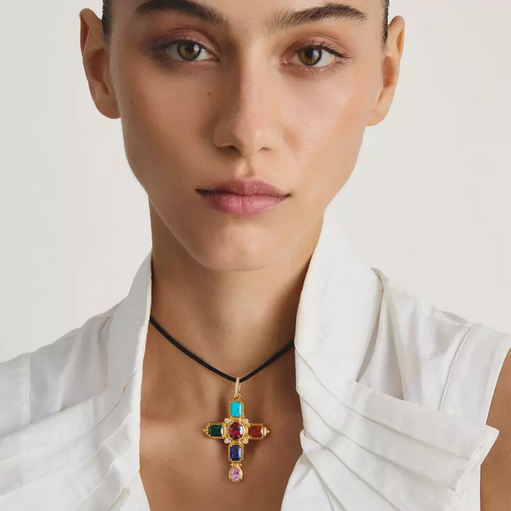A close-up of a model wearing a gold cross pendant necklace with colorful gemstone accents on a black cord, paired with a white shirt.