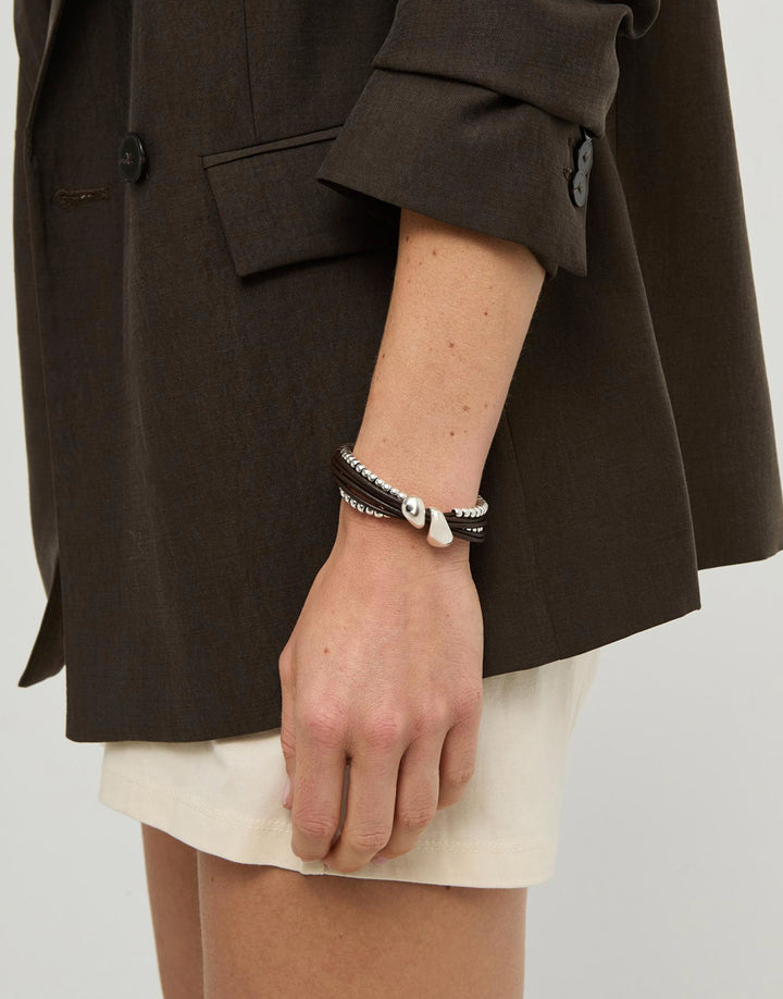 A close-up of a person's hand and wrist wearing a multi-strand bracelet with black cords, silver-toned metal, and a small white bead charm, styled with a dark brown blazer and cream shorts.