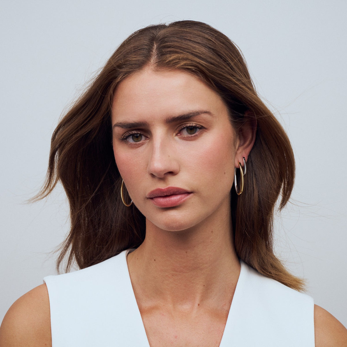 Woman with long brown hair wearing gold hoop earrings against a light gray background