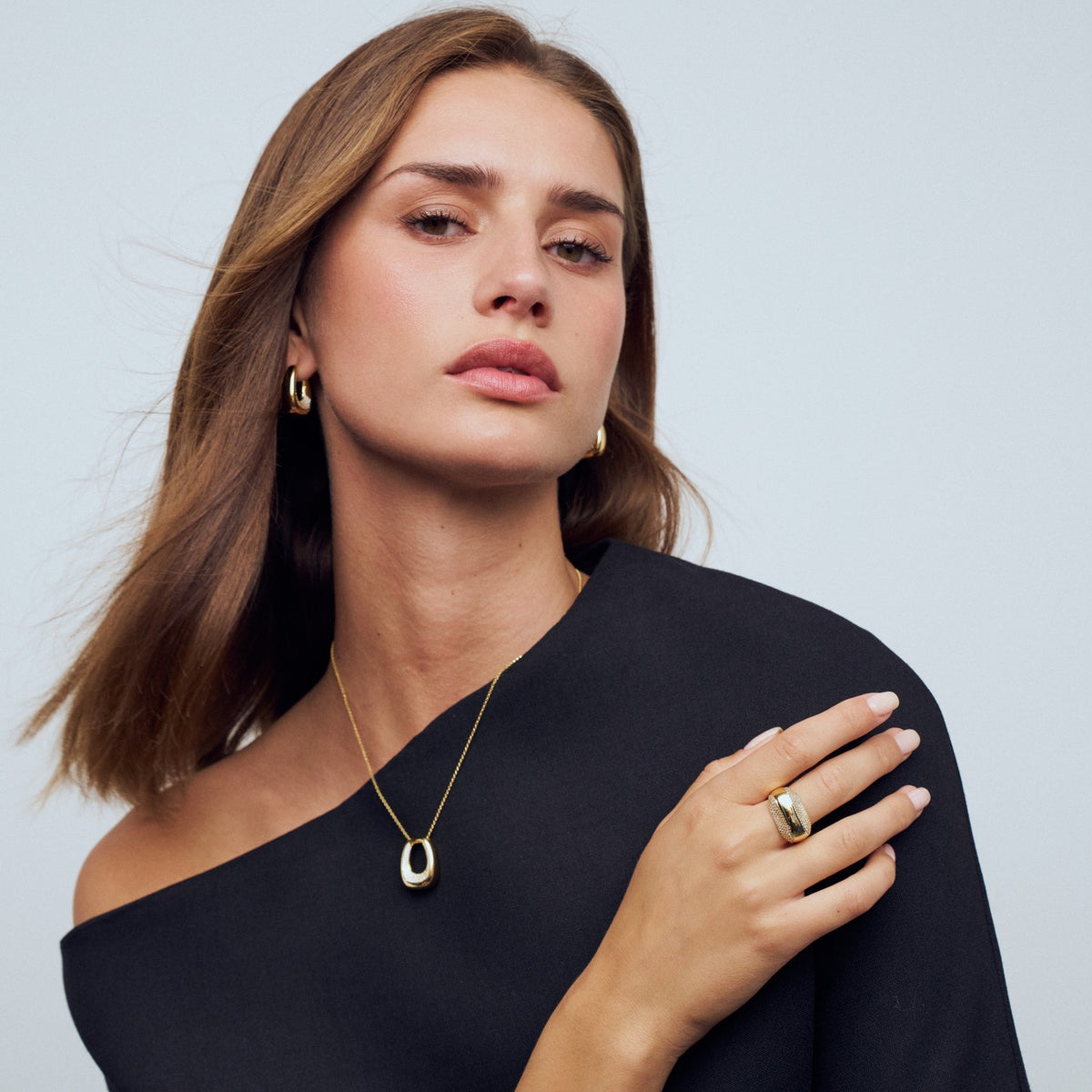 Woman wearing gold jewelry including a necklace, earrings, and ring against a light gray background