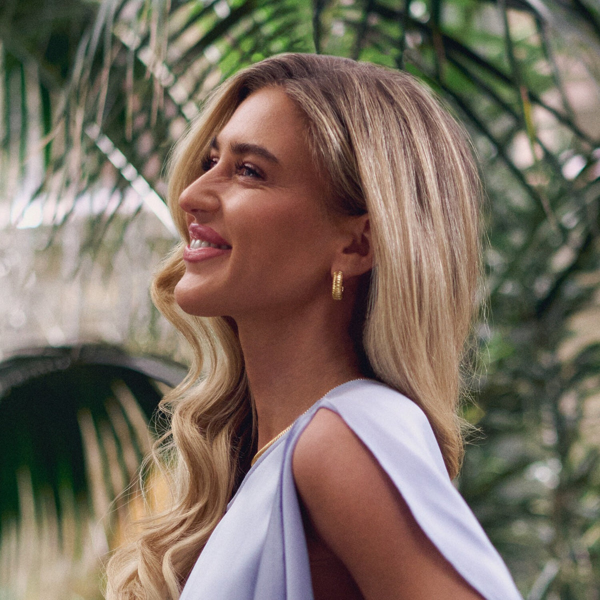 Woman with blonde hair and a light blue top standing among green foliage.