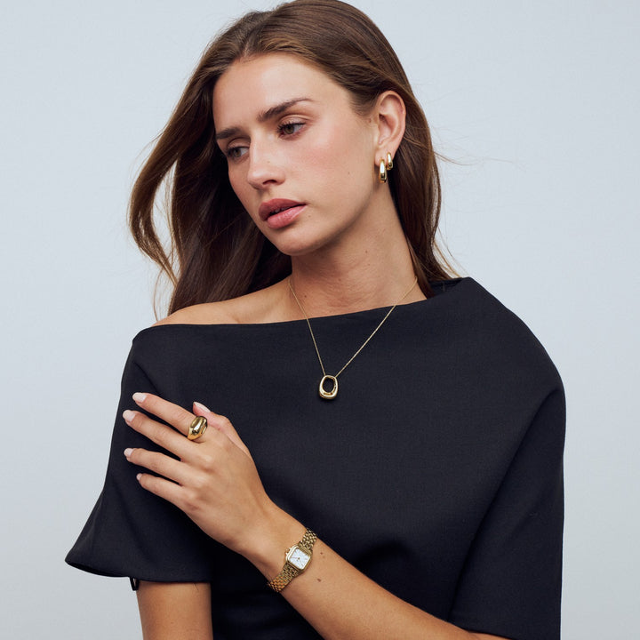 Woman wearing gold jewelry including a necklace, ring, and bracelet against a light gray background