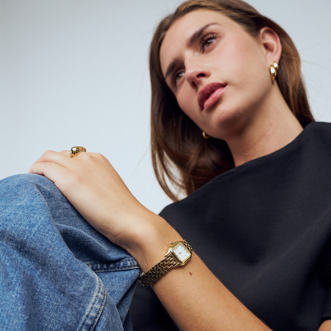 Woman wearing a gold watch and ring, sitting with a neutral background