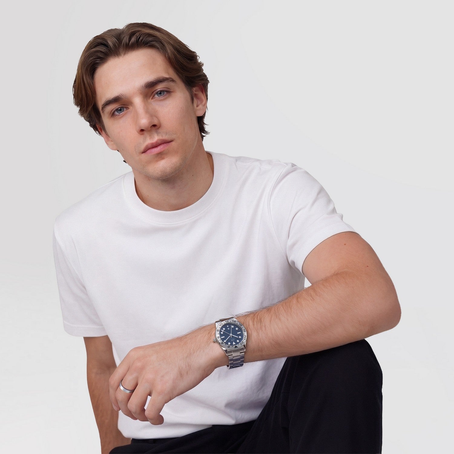 Man wearing a white t-shirt and dark pants with a watch on a light gray background