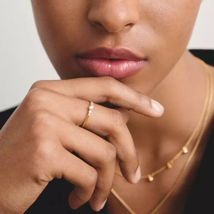 A close-up lifestyle shot of a model wearing a delicate gold ring adorned with three small white pearls on her ring finger, paired with a gold necklace.