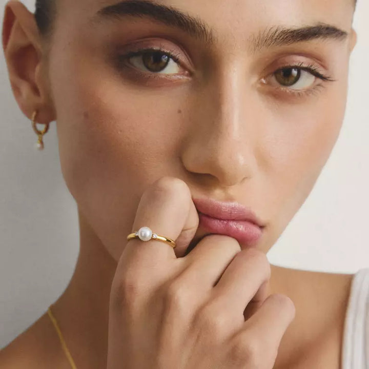 A close-up lifestyle shot of a model wearing a delicate gold ring with a single white pearl on her finger, set against a soft, light neutral background.